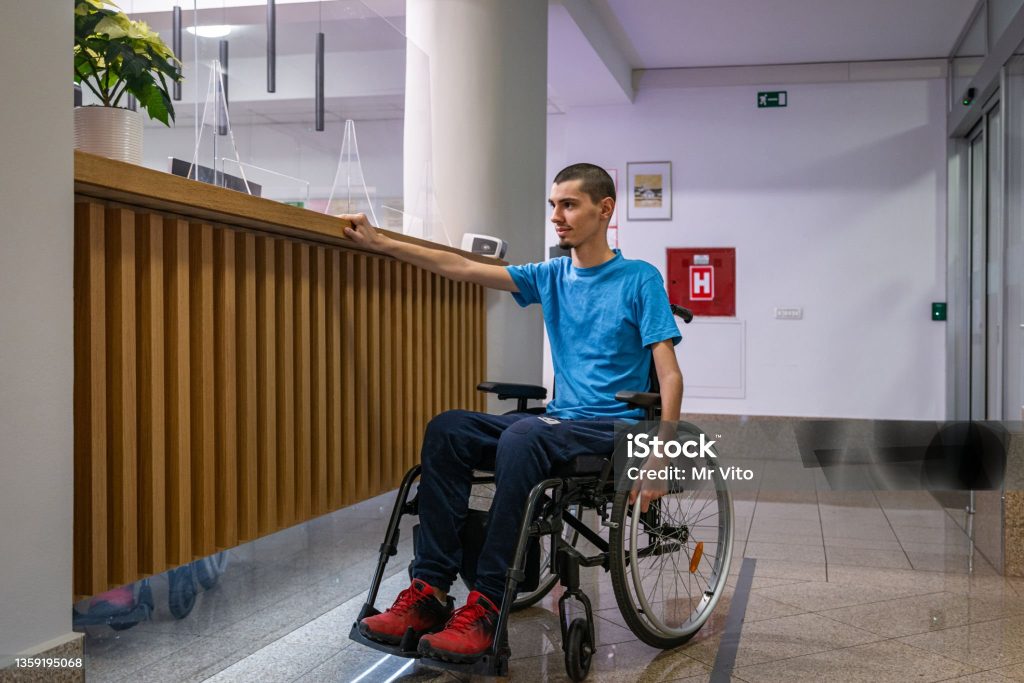 A disabled teenager with dystrophy in a blue T-shirt in a wheelchair stands in front of a too-high counter at the hotel reception.